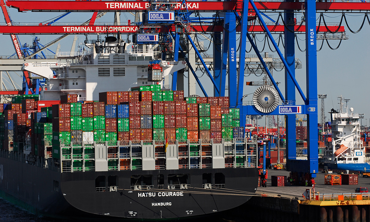 schwarzes Containerschiff liegt unter den blau-roten Kraenen am Terminal Burchardkai im Hafen von Hamburg