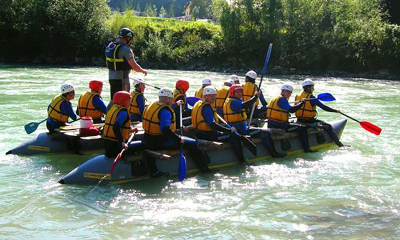 Eine mit Rettungswesten und Helmen ausgestattete Gruppe paddelt auf einem Floss ueber einen Fluss