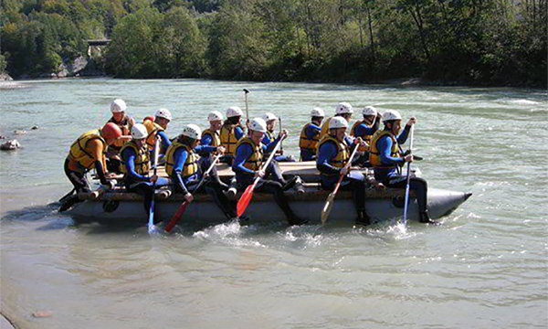 Eine Veranstalungsgruppe paddelt, ausgestattet mit Rettungswesten und Helmen, ein Floss ueber einen Fluss